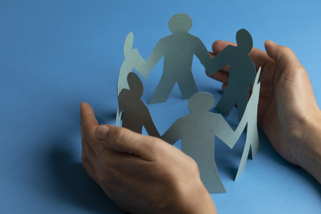 Paper people surrounded by hands in gesture of protection on blue background.